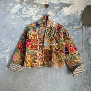 May include: A quilted jacket featuring a patchwork design of various floral and botanical patterns in shades of tan, red, blue, and green. The jacket has long sleeves with striped cuffs, a front opening, and white stitched trim along the edges.