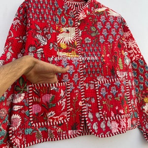 May include: A red quilted jacket with a floral print and a mandarin collar. The jacket has a button closure and a pocket on the left side.