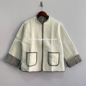 May include: Cream quilted jacket with black and white striped detailing on the collar, cuffs, and button line. It has two front pockets with black trim. The jacket has a stand-up collar and long sleeves. The text "fashionandfurnish" is visible on the jacket.