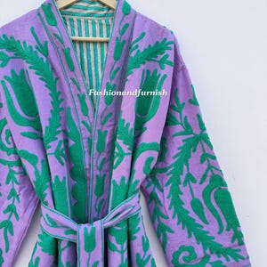 May include: A lilac dressing gown with green floral embroidery and a matching belt. The interior of the gown has a teal and white striped pattern. The text "Fashionandfurnish" is visible. The gown is hanging on a wooden hanger.