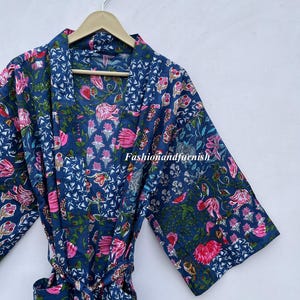 May include: A blue floral print robe with a matching belt, hanging on a wooden hanger. The robe features a vibrant pattern of pink flowers and green leaves against a dark blue background. The text "Fashionandfurnish" is visible.
