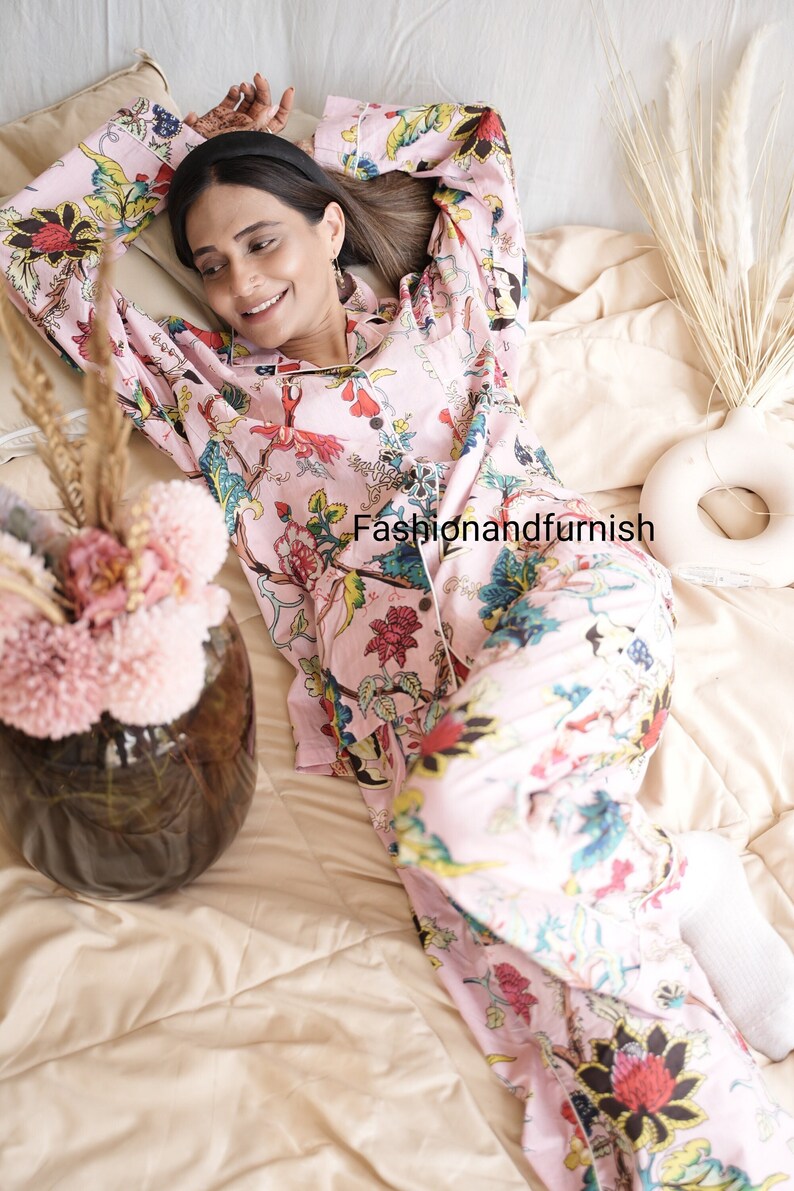 May include: A light pink pajama set featuring a vibrant floral pattern with red, yellow, blue, and green botanical motifs. The long-sleeved button-down top and matching trousers include the text Fashionandfurnish printed on the fabric.