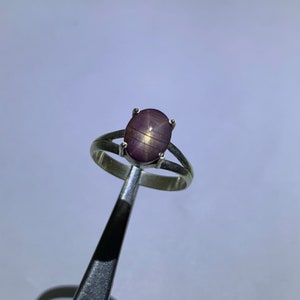 May include: A silver ring featuring a purple star ruby gemstone. The ruby has a star-shaped pattern in the centre.