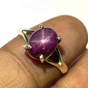 May include: A silver ring with a purple star ruby gemstone. The ruby is oval shaped and has a star-like pattern.
