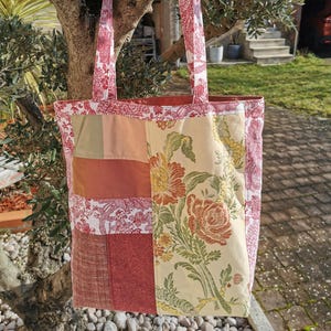 Op de afbeelding: Een patchwork tote bag met een bloemenprint. De tas is gemaakt van verschillende stoffen in tinten rood, roze, groen en beige. De tas heeft twee hengsels en een groot hoofdvak.