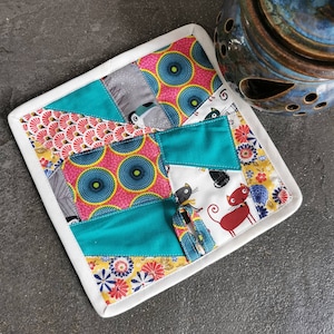 Op de afbeelding: Een witte, gestikte onderzetter met een patchwork-ontwerp met turquoise, roze, gele en zwarte stof. De stof heeft verschillende patronen, waaronder cirkels, bloemen en dieren.