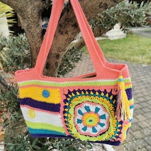 Peut inclure: Un sac fourre-tout en crochet avec des rayures colorées et un grand motif de fleur en crochet. Le sac a deux anses roses et un pompon jaune.