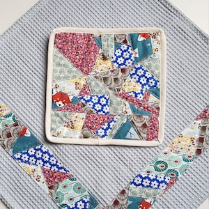 Peut inclure: Une serviette de cuisine en tissu gaufré gris avec un dessous de plat de style patchwork. Le dessous de plat est fait de différents tissus avec des motifs floraux et géométriques dans des tons de rose, bleu, vert et jaune. Le dessous de plat a une bordure de couleur crème.