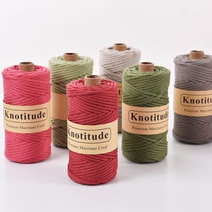 Puede incluir: Seis bobinas de cordón macramé premium Knotitude en varios colores, incluyendo rojo, verde, gris y marrón. Cada bobina tiene un núcleo de cartón marrón y una etiqueta con el nombre de la marca y la descripción del producto. Los cordones están bien enrollados.