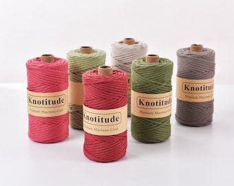 May include: Six spools of Knotitude premium macrame cord in various colors, including red, green, gray, and brown. Each spool has a brown cardboard core and a label with the brand name and product description. The cords are tightly wound.