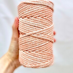 Puede incluir: Un carrete de cordón de macramé en una mezcla de melocotón y blanco. El cordón está bien enrollado, creando una superficie texturizada. La combinación de colores da una estética suave y neutra, adecuada para varios proyectos de artesanía. El cordón es sostenido por una mano.