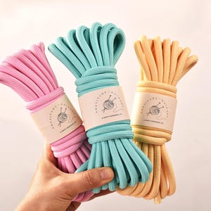 Puede incluir: Tres ovillos de cordón de macramé de colores rosa, turquesa y amarillo. Cada ovillo está envuelto con una etiqueta con un logotipo de agujas e hilo. El cordón está bien enrollado, lo que sugiere que está listo para proyectos de manualidades.