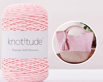 Puede incluir: Una bobina grande de hilo de macramé suave de poliéster rosa con la marca "knotitude" en la etiqueta. Un bolso tote rosa y beige tejido a ganchillo con una borla está en un círculo.