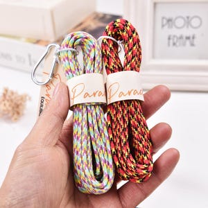 May include: Two colorful paracord ropes, one multi-colored and the other red, yellow, and black, are held in a hand. Each rope is wrapped with a paper band that reads "Para Para". A silver carabiner clip is attached to the multi-colored rope.