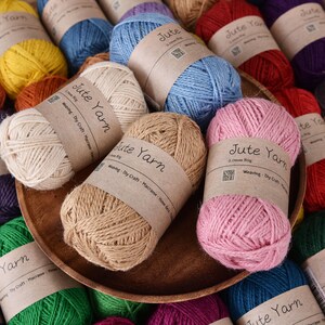 May include: A collection of jute yarn skeins in various colors, including pink, blue, yellow, and green. Each skein is wrapped with a brown paper label that reads "Jute Yarn". The yarn is arranged in a wooden bowl and scattered around it.