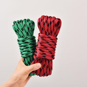 May include: Two coiled ropes, one green and one red, each with a black diamond pattern. The ropes are held against a white background, showcasing their texture and color contrast.