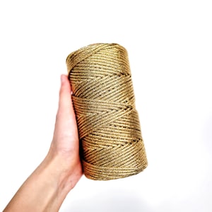 May include: A spool of gold metallic ribbon. The ribbon is tightly wound and has a textured surface. The gold color has a slight shimmer, reflecting light. The ribbon is held in a hand against a white background.