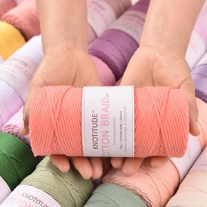 Könnte beinhalten: Eine Spule korallenfarbenes Baumwoll-Zopf-Garn wird von Händen gehalten. Das Etikett lautet "KNOTITUDE COTTON BRAID 20". Im Hintergrund sind weitere Garnspulen in verschiedenen Farben, darunter Grün, Orange und Lila, zu sehen.