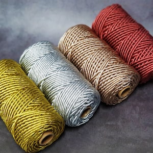 May include: Four spools of decorative twine in a row, each a different colour. The colours are gold, silver, bronze, and red. The twine is tightly wound around a cardboard core, and the background is a dark grey.