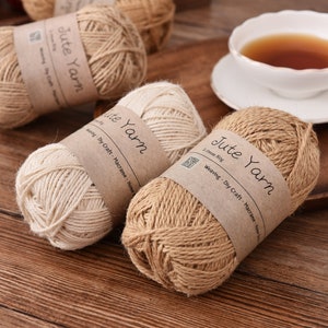 May include: Several spools of jute yarn in varying shades of beige and brown are displayed. Each spool has a brown paper label that reads "Jute Yarn". A white teacup with tea sits on a wooden tray, suggesting a craft or home decor theme.