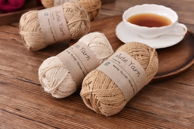 May include: Several spools of jute yarn in varying shades of beige and brown are displayed. Each spool has a brown paper label that reads "Jute Yarn". A white teacup with tea sits on a wooden tray, suggesting a craft or home decor theme.