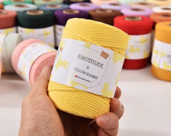 May include: A hand holds a yellow spool of cotton cord with a white label that reads "KNOTITUDE x YELLOW RABBIT" with a yellow rabbit graphic. Other spools of various colors are in the background.