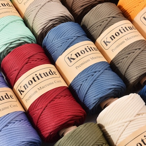 May include: A selection of macrame cord spools in various colours. The spools are labelled "Knotitude Premium Macrame Cord". Colours include blue, red, green, and orange. The cord is likely sold by the metre.