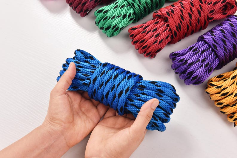 May include: A close-up of several colorful ropes. A blue rope with black accents is held in the center. Other ropes in red, green, purple, and yellow are visible in the background. The ropes are tightly coiled.