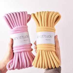May include: Two bundles of macrame cord in pink and yellow are held up against a white background. Each bundle is wrapped with a white label that reads "knotitude" and "Macrame". The cord is thick and tightly wound.