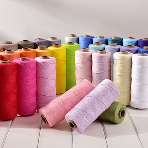 Pode incluir: Uma coleção de rolos de barbante coloridos em vários tons, incluindo vermelho, rosa, amarelo, verde, azul e branco. Os rolos estão dispostos sobre uma superfície de madeira branca, mostrando as cores vibrantes e as texturas do barbante.