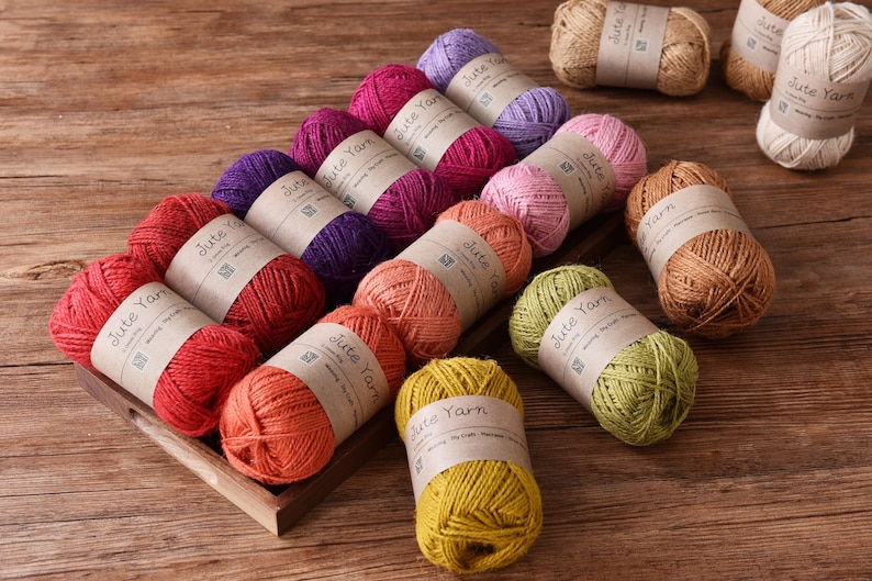 May include: A collection of colorful jute yarn skeins in shades of red, orange, pink, purple, green, and yellow. Each skein is labeled with a brown paper band that reads "Jute Yarn". The yarn is arranged on a wooden tray.