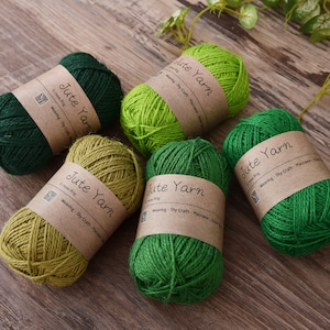 May include: Five skeins of jute yarn in shades of green and olive, each wrapped with a brown paper label. The labels read "Jute Yarn" and include text about weaving and DIY crafts. The yarn is arranged on a wooden surface.