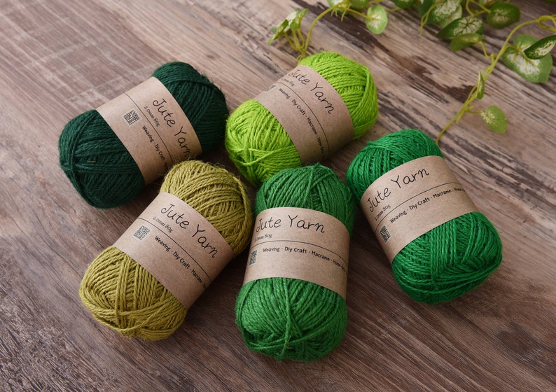 May include: Five skeins of jute yarn in shades of green and olive, each wrapped with a brown paper label. The labels read "Jute Yarn" and include text about weaving and DIY crafts. The yarn is arranged on a wooden surface.