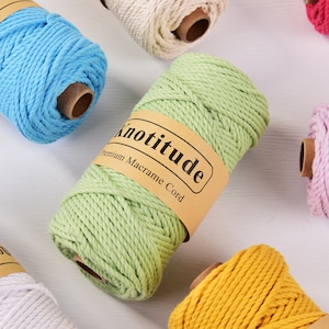 Könnte beinhalten: Eine Auswahl an farbigen Makramee-Kordelspulen in verschiedenen Farbtönen, darunter Blau, Grün, Gelb, Rosa und Weiß. Jede Spule hat ein braunes Papieretikett mit dem Text "Knotitude Premium Macrame Cord". Die Spulen sind auf einer weißen Oberfläche angeordnet.