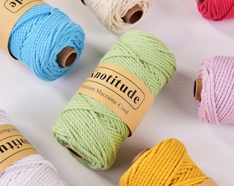 May include: A collection of colorful macrame cord spools in various shades, including blue, green, yellow, pink, and white. Each spool has a brown paper label with the text "Knotitude Premium Macrame Cord". The spools are arranged on a white surface.