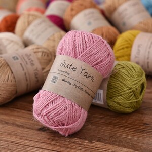 May include: A collection of jute yarn balls in various colors, including pink, yellow, and natural tones. Each ball is wrapped with a brown paper label that reads "Jute Yarn". The yarn is suitable for weaving, dry craft, and macrame projects.