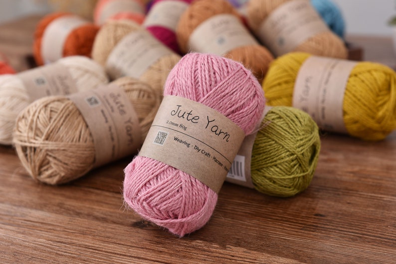 May include: A collection of jute yarn balls in various colors, including pink, yellow, and natural tones. Each ball is wrapped with a brown paper label that reads "Jute Yarn". The yarn is suitable for weaving, dry craft, and macrame projects.