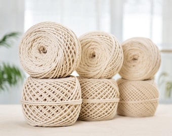 May include: Six beige yarn balls are stacked on a light-colored surface. The yarn is tightly wound, creating a textured appearance. The balls are arranged in a visually appealing composition, suggesting a craft or DIY project.