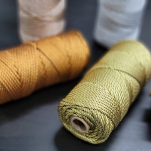 May include: Close-up of several spools of colourful macrame cord. The cord is in shades of gold, green, and pink, with other spools in the background. The cord is tightly wound on cardboard spools, ready for crafting projects.