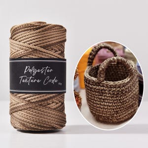 Op de afbeelding: Een spoel bruin polyestergaren met een zwart etiket met de tekst "Polyester Texture Code". Er is ook een kleine, handgehaakte bruine mand met een handvat te zien.
