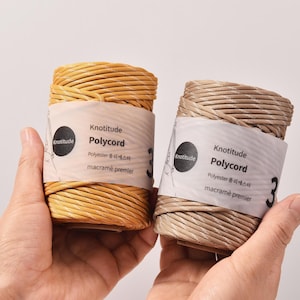 Puede incluir: Dos bobinas de cordón Polycord para macramé en tonos beige y marrón claro se sostienen en las manos. Cada bobina tiene una etiqueta con el logotipo de Knotitude e información del producto. El cordón está bien enrollado.