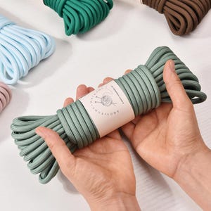 Op de afbeelding: Een streng saliegroen macramékoord in handen gehouden, met een label met de tekst "KNOTITUDE". Andere strengen koord in verschillende kleuren, waaronder blauw, roze, groen, bruin, geel en paars, zijn op de achtergrond zichtbaar.