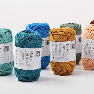 May include: A collection of colorful Daisy Yarn skeins in shades of teal, blue, mustard yellow, peach, green, and brown. Each skein is wrapped with a label that reads "Daisy Yarn" and features a heart design. The yarn is tightly wound, suggesting a durable texture.