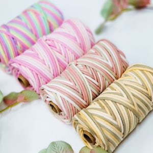 May include: Four spools of colorful macrame cord in pastel shades of pink, yellow, green, and purple. The cord is tightly wound on cardboard spools, perfect for crafting projects. The cords are arranged diagonally, with green and pink leaves in the background.