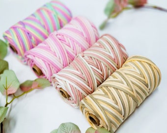 May include: Four spools of colorful macrame cord in pastel shades of pink, yellow, green, and purple. The cord is tightly wound on cardboard spools, perfect for crafting projects. The cords are arranged diagonally, with green and pink leaves in the background.
