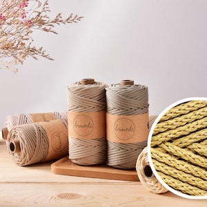 Puede incluir: Se muestran bobinas de cordón de macramé beige sobre una superficie de madera, algunas con etiquetas marrones que dicen "brownie". Un primer plano revela la textura tejida del cordón. Flores secas en una cesta de mimbre en el fondo.