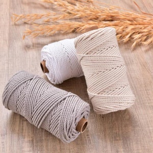 Può includere: Tre rocchetti di cordoncino per macramè bianco, beige e grigio, disposti su una superficie di legno chiaro. Il cordoncino è ben avvolto, mostrando la consistenza del materiale. Spighe di grano secche sullo sfondo.