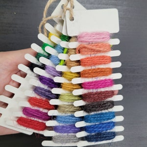 May include: A color swatch of various shades of yarn. The yarn is arranged on a white plastic holder with blank tags. The colors include pink, orange, red, blue, and green. The holder is tied with twine.