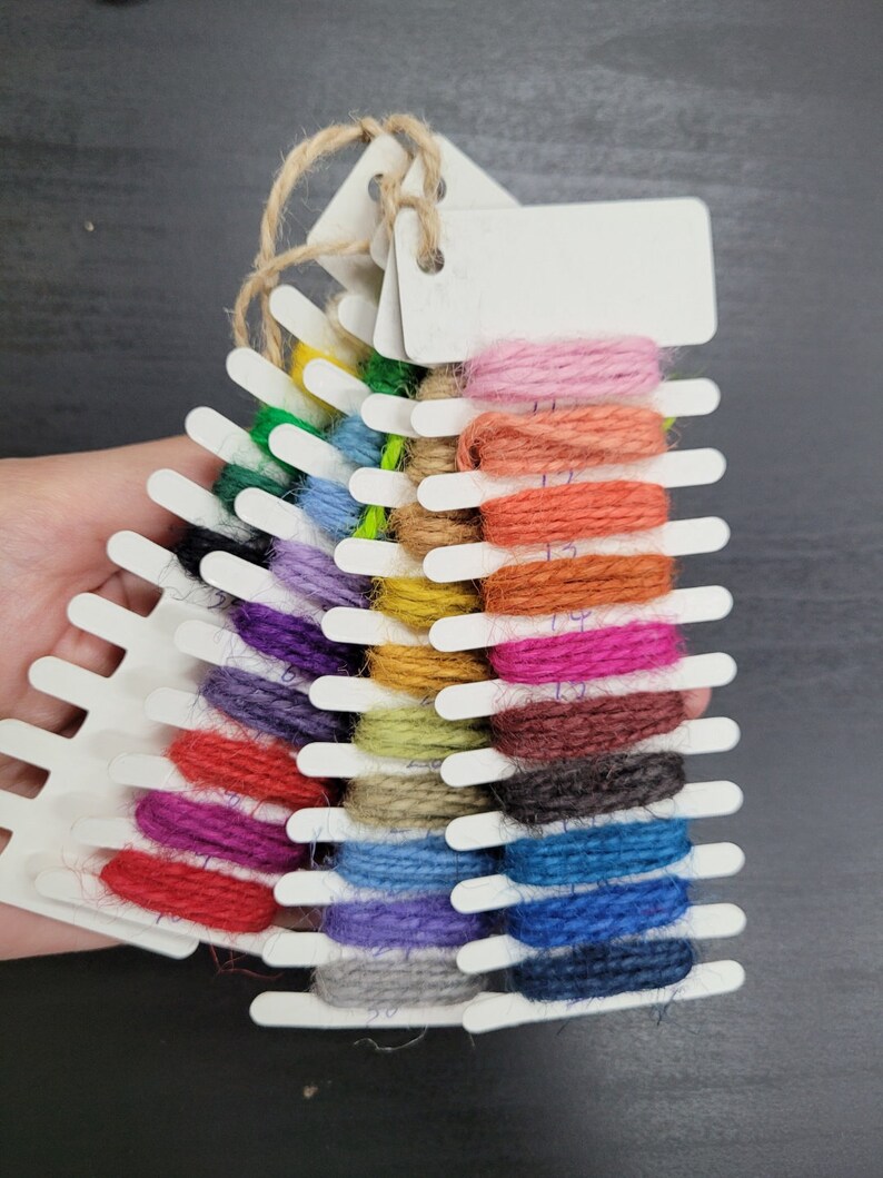 May include: A color swatch of various shades of yarn. The yarn is arranged on a white plastic holder with blank tags. The colors include pink, orange, red, blue, and green. The holder is tied with twine.