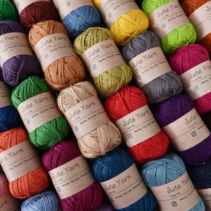 May include: A collection of colorful jute yarn balls, each wrapped with a brown paper label that reads "Jute Yarn". The yarn is available in various colors, including green, red, blue, and brown. Ideal for weaving, dry craft, and macrame projects.
